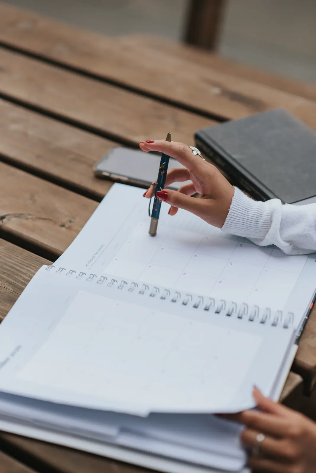 A hand holding a pen over an open notebook on a wooden table outdoors.