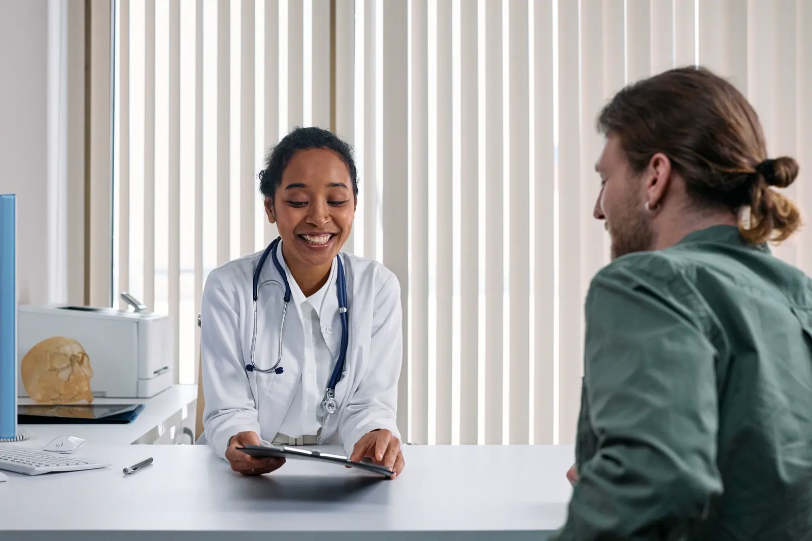 A clinician in a white coat speaking with a patient in a bright exam room.
